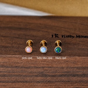 May include: Three gold-tone stud earrings with round cabochon stones. The left earring features a pink opal, the center earring has a baby blue opal, and the right earring contains a black opal. Text below each stone reads pink opal, baby blue opal, and black opal.