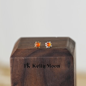 Puede incluir: Un par de pendientes de botón con centros de piedras preciosas naranjas. Un pendiente tiene un engaste dorado, el otro plateado. Los pendientes se exhiben en un bloque de madera oscura con el texto "1R Kelly Moon".