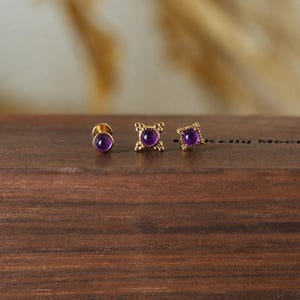 Könnte beinhalten: Drei goldfarbene Ohrstecker mit lila Edelsteinen. Die Ohrringe werden auf einer dunklen Holzoberfläche präsentiert. Ein Ohrring ist rund, einer ist quadratisch und einer hat ein dekoratives Kreuzdesign.