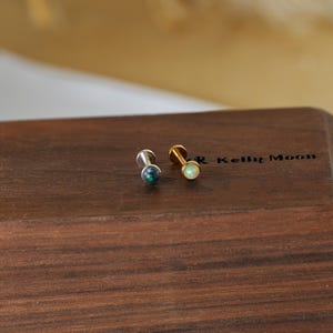 May include: Two small stud earrings. One is silver with a dark blue and green stone, and the other is gold with a white stone. The earrings are displayed on a dark wooden surface.