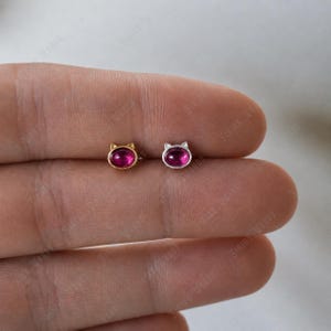 以下が含まれることがあります： 猫の形をしたスタッドピアス2つ。片方は金色、もう片方は銀色です。どちらも鮮やかな楕円形のマゼンタ色の石が中央にセットされています。ピアスは小さく繊細で、普段使いに最適です。
