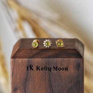 Puede incluir: Tres pendientes de oro con piedras preciosas verde claro se exhiben sobre un bloque de madera. Los pendientes tienen diferentes diseños: una piedra redonda, una forma de flor con piedras claras y una forma cuadrada con un borde de cuentas. El bloque está grabado con el texto "1R Kelly Moon."