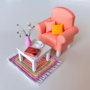 May include: Miniature living room set with a coral armchair, two pink pillows, and an orange pillow. A white table holds a book, a cup, and a pink vase with flowers. A colorful rug completes the scene.