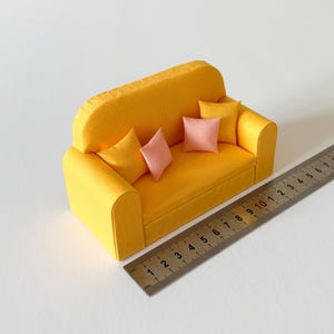 Może przedstawiać: Miniaturowa żółta sofa z trzema małymi, jasnoróżowymi kwadratowymi poduszkami dekoracyjnymi. Metalowa linijka umieszczona wzdłuż przedniej podstawy pokazuje, że długość sofy wynosi około 10,5 centymetra. Sofa posiada oparcie i dwa podłokietniki.