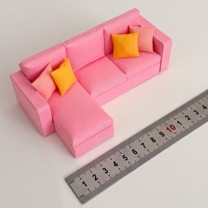 May include: A miniature pink sectional sofa with three throw pillows in shades of orange and pink. A metal ruler is placed next to the sofa, showing measurements in millimeters.