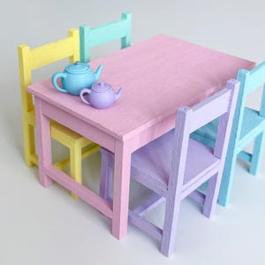 May include: A miniature wooden dining set featuring a rectangular pink table and four chairs in pastel yellow, mint green, light blue, and lavender. Two small ceramic teapots, one light blue and one lavender, are placed on the tabletop surface.