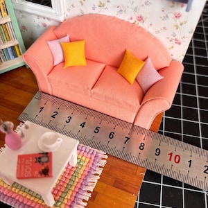 Könnte beinhalten: Miniatur-Couch in Pfirsichfarbe mit drei gelben und rosa Kissen. Ein kleiner weißer Tisch mit einem Buch und einem bunten Teppich befindet sich vor der Couch. Die Couch ist etwa 10 cm lang, wie das Lineal zeigt.