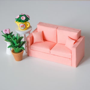 Könnte beinhalten: Ein Miniatur-Puppenhaus-Wohnzimmer-Set mit einem rosa Sofa mit zwei passenden Kissen, einem kleinen weißen Tisch mit einer gelben Teekanne mit Blumen und mehreren Topfpflanzen. Das Set ist in einer Pastellfarbpalette gestaltet.
