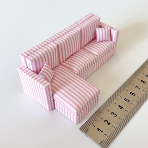 May include: A miniature L-shaped sofa with a pink and white striped pattern. The sofa includes two small matching pillows. A ruler is placed next to the sofa for scale.