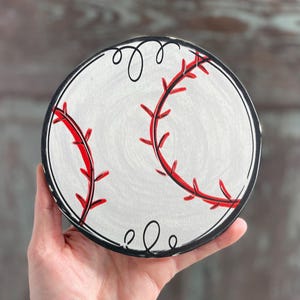 May include: A white wooden round sign with a black outline and red stitching, depicting a baseball.