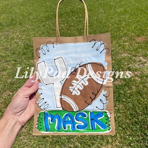 May include: A brown paper gift bag with a football-themed design. The bag features a painted football, goalpost, and the word "MASK" in blue and green. The bag has a brown twisted paper handle.