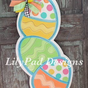 May include: A wooden Easter door hanger with three stacked eggs. The top egg is yellow with wavy lines, the middle egg is green with wavy lines, and the bottom egg is orange with wavy lines. Each egg has a blue outline and is decorated with colorful polka dots.