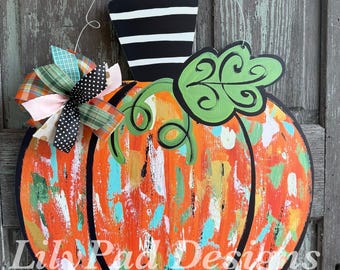 May include: A pumpkin-shaped wooden door hanger featuring a black and white striped stem, a green leaf, and a multi-colored painted surface with orange, white, and teal brushstrokes. A decorative bow with plaid and polka dot ribbons is attached.