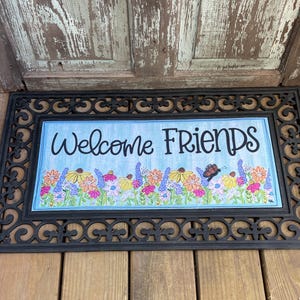 May include: A black welcome mat with an ornate metal frame and a blue background. The mat has a floral design with a butterfly and the text "Welcome Friends".