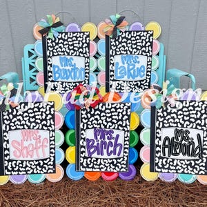 May include: Wooden teacher door hangers shaped like composition notebooks with the names "Mrs. Buxton", "Mrs. LaRue", "Mrs. Shaff", "Mrs. Birch", and "Ms. Almond". Each notebook is set against a colorful, layered background and topped with a decorative bow.