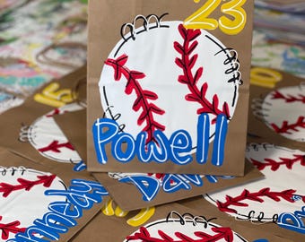 May include: A brown paper gift bag featuring a white baseball graphic with red stitching, the yellow number 23 at the top, and the blue name Powell printed across the center. The bag has brown paper handles and a flat rectangular base.