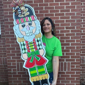 May include: A large, colorful wooden nutcracker decoration. The nutcracker has a black hat with a striped band, a red and green outfit, and a large bow at the top. The background is a brick wall.