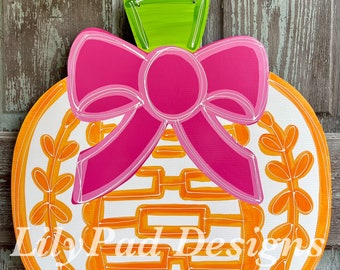 May include: A wooden door hanger in the shape of a pumpkin with a bright pink bow and green top. The pumpkin is white with orange accents and a decorative design. The text "Lily Pad Designs" is visible.