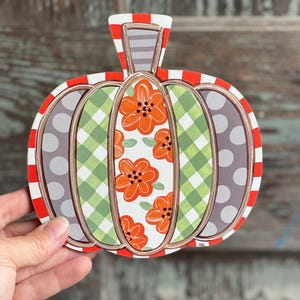 May include: A decorative wooden pumpkin with a red and white striped border. The pumpkin features sections of gray polka dots, green gingham, and orange floral patterns. The stem is striped gray and white.