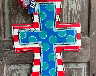 May include: A wooden cross painted in red, white, and blue with a blue center featuring white polka dots. A red, white, and blue bow is attached to the top of the cross.