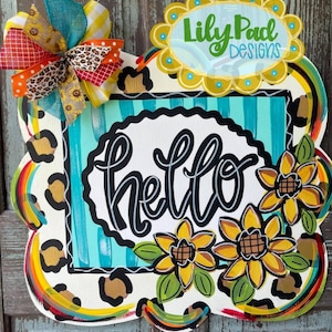 Puede incluir: Un letrero de bienvenida para la puerta con un fondo de estampado de leopardo, un cuadrado a rayas turquesa y blanco con la palabra "hello" en letra negra, y tres girasoles amarillos con hojas verdes.