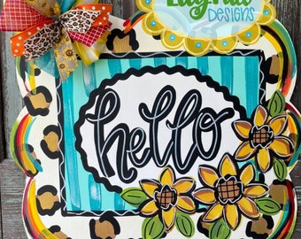 May include: A welcome door sign with a leopard print background, a turquoise and white striped square with the word "hello" in black script, and three yellow sunflowers with green leaves.