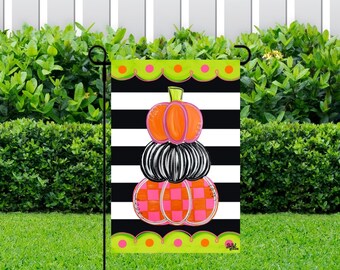 May include: A black and white striped garden flag featuring a stack of three pumpkins. The top pumpkin is orange with pink accents, the middle pumpkin is black with white stripes, and the bottom pumpkin is pink with orange and white checkered accents. The flag has a green border with pink and orange polka dots.
