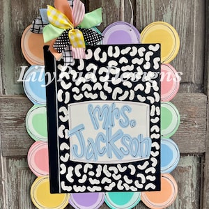 May include: A colorful teacher-themed door hanger featuring a black notebook design with white spots and the name "Mrs. Jackson" in blue. The door hanger is surrounded by pastel-colored circles and topped with a multi-colored bow.