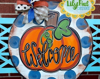 May include: Round wooden welcome sign featuring a hand-painted orange pumpkin with green leaves and the word "Welcome" in black script. The sign has blue polka dots and a decorative bow.
