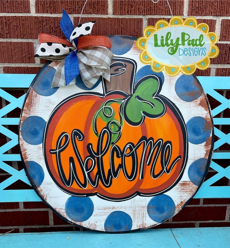 May include: A round wooden door hanger with a white background and blue polka dots. A painted orange pumpkin with a brown stem and green leaves is in the center of the door hanger. The words 'Welcome' are written in orange cursive lettering on the pumpkin. The door hanger has a brown and white plaid bow and a blue and white polka dot bow attached to the top.