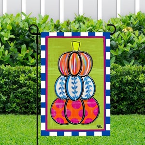 May include: A colorful, hand-painted stack of pumpkins with blue, orange, pink, and white accents. The pumpkins are on a green background with a blue and white checkered border.