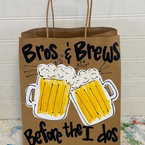 May include: A brown paper gift bag with black handles. The bag is decorated with a hand-drawn design of two beer mugs with the text "Bros & Brews Before the I Dos" in black.