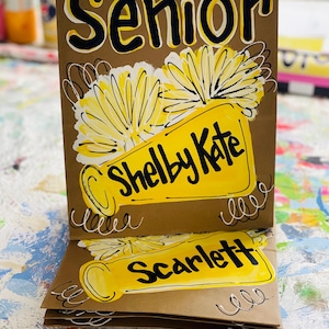 May include: Three brown paper gift bags with yellow and black painted designs. The top bag has the word "Senior" written in black with yellow outlines. The middle bag has a yellow megaphone with the words "Shelby Kate" written in black. The bottom bag has a yellow megaphone with the word "Scarlett" written in black.