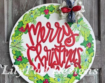 May include: A round, white Christmas door hanger with the words "Merry Christmas" in red script. The words are surrounded by a green and red holly wreath. A decorative bow with red and blue accents is attached at the top.