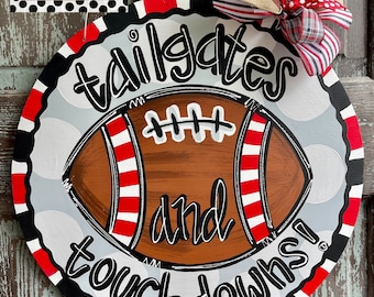 Peut inclure: Une porte ronde en bois avec un ballon de football peint et les mots "tailgates and touchdowns!" en noir et blanc. Le ballon de football a des rayures rouges et blanches. La porte est décorée d'un nœud rouge, blanc et noir.