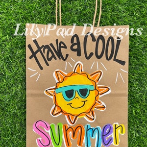 May include: A brown paper gift bag with a white handle. The bag is decorated with black marker writing that says "Have a Cool Summer". A yellow sun with sunglasses is drawn in the center of the bag. The word "Summer" is written in rainbow colors.