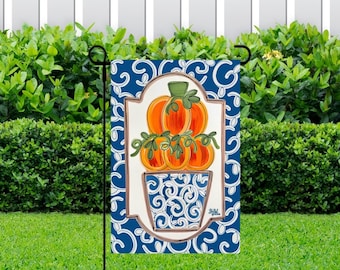May include: A blue and white garden flag with a white frame and a design of orange pumpkins in a blue and white pot with a white swirl pattern. The pumpkins are stacked on top of each other and have green stems.