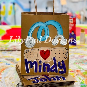 May include: Brown paper gift bags with blue and white painted bandaids, a red heart, and the names Mindy and John in purple.