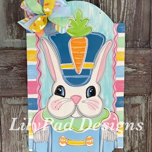 May include: A painted wooden door hanger featuring a cartoon bunny wearing a blue and yellow drum major uniform with a carrot on top. The bunny has large eyes and a pink nose. The background is a blue and white striped pattern with a pink and yellow bow at the top.