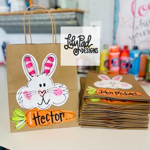 May include: Brown paper gift bags decorated with a white bunny face and a carrot. The carrot has the name "Hector" written on it. The bags are stacked on top of each other. The top bag has the name "John Michael" written on it.