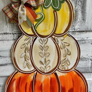 May include: A stack of three pumpkins painted in fall colors. The top pumpkin is yellow with a green stem and leaves. The middle pumpkin is beige with brown vines. The bottom pumpkin is orange with brown lines. A brown and gold bow is tied around the top pumpkin.