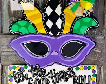 May include: A purple and gold Mardi Gras door hanger with a jester's hat, mask, and beads. The sign reads "Let the good times roll."