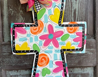 May include: A colorful wooden cross with a floral design painted on it. The cross is decorated with a bright pink, yellow, and black bow. The cross is hanging on a wooden door. The text "Lily Pad Designs" is visible in the bottom right corner of the image.
