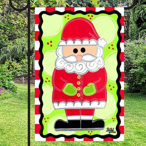 May include: A Christmas garden flag featuring a cartoon Santa Claus design. Santa is depicted in a red suit and hat with a white beard and green mittens. The flag has a green background with white polka dots and a red and white striped border.