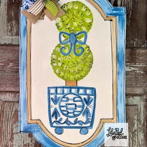 May include: A hand-painted wooden sign with a blue and white topiary design. The sign features a green topiary with a blue bow in a blue and white pot. The sign has a blue border and a gold accent. The sign is signed "Lily Pad Designs".
