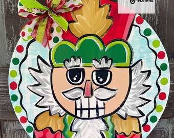 May include: A hand-painted wooden nutcracker door hanger with a red, green, and white color scheme. The nutcracker has a green hat, a white beard, and a red and white striped uniform. The door hanger is decorated with a red and green polka dot bow.