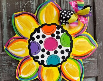 May include: A large, yellow sunflower door hanger with a black and white polka dot center. The center has colorful circles in shades of pink, purple, green, orange, and blue. A rainbow and black and white polka dot bow is attached to the top of the sunflower.