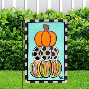 May include: A colorful fall-themed garden flag with a stack of pumpkins. The top pumpkin is orange, the next is brown with black polka dots, and the bottom is a mix of yellow, green, and pink stripes. The flag has a black and white checkered border.
