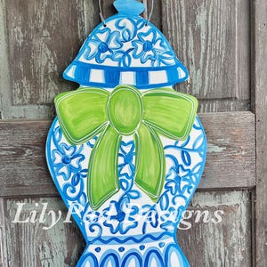 May include: A wooden decorative vase in blue and white with a green bow. The vase has a lid and a beaded handle. The design includes floral patterns and stripes. The text "Lily Rose Designs" is visible.