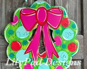 May include: A wooden wreath with a large pink bow and colourful polka dots. The wreath has a green background with red, blue, and pink polka dots. The text "Lily Pad Designs" is visible.